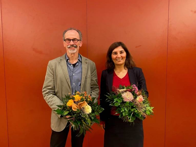 Ein Mann und eine Frau stehen vor einer roten Wand und halten jeweils einen Blumenstrauß in den Händen.